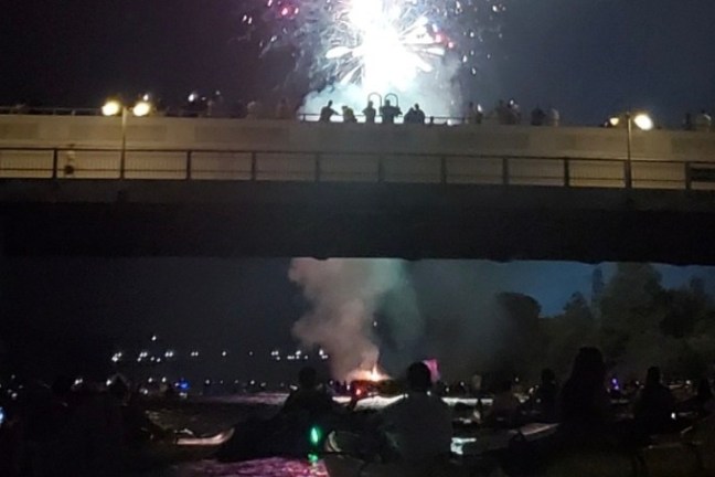 People in kayaks on a river at night watching fireworks above a bridge.